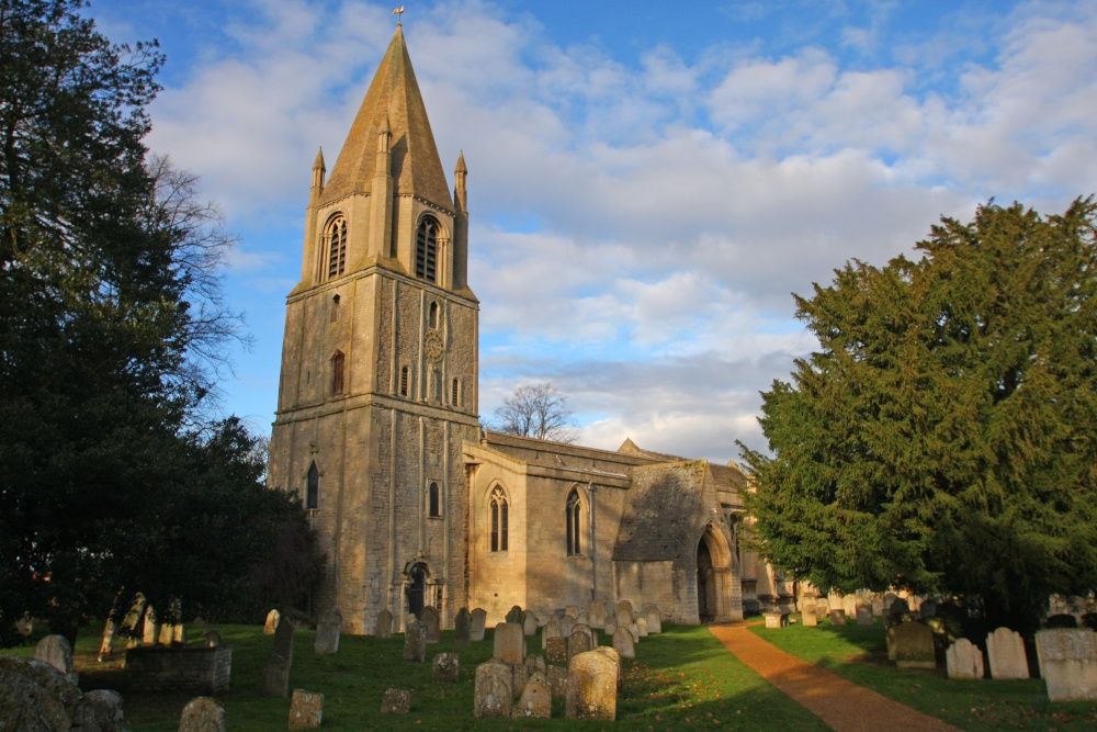 A view of Barnack Church surrounded by trees under white clouded and blue skies